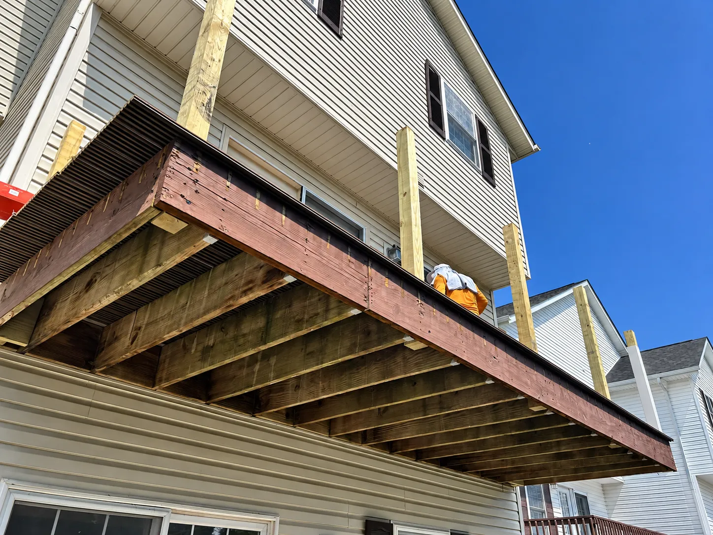 Underside view of an elevated residential deck during construction, showing wooden floor joists, support beams, and new railing posts.